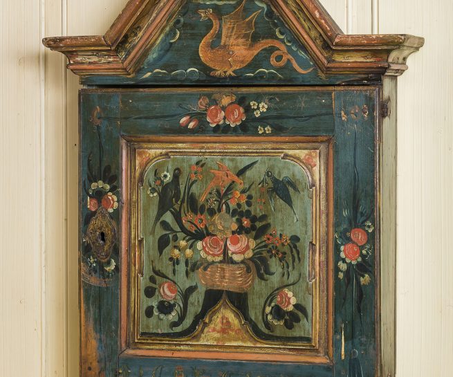 Hengeskap fra Telemark datert 1792, med malte roser, fugler og en drage. Et unikt arbeid av Olav Hansson. Speilet i døren er skåret og fargene vakkert avstemt til hverandre. Det er påmalt et dobbelt sett initialer og årstall. Telemark og svenskeskap hadde døra over hele fronten.